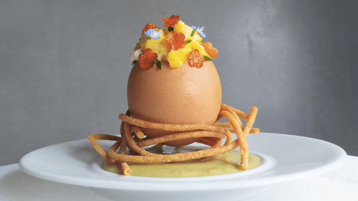Un nido di spaghetti croccanti con un guscio d'uovo ripieno di uova strapazzate al timo e vellutata di asparagi. A decorare tutto dei deliziosi fiorellini di carote saltate in padella.