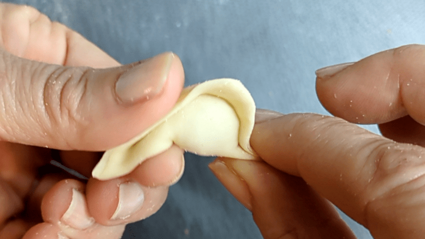 Penultimo passo per fare un tortellino, la piega sulla parte sinistra del triangolino di pasta con ripieno.