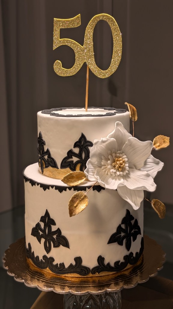 Una elegantissima torta per i 50 anni di una splendida fanciulla: base bianca e ora, decorazioni in pizzo di zucchero nere e una splendida camelia fatta a mano a guarnizione.